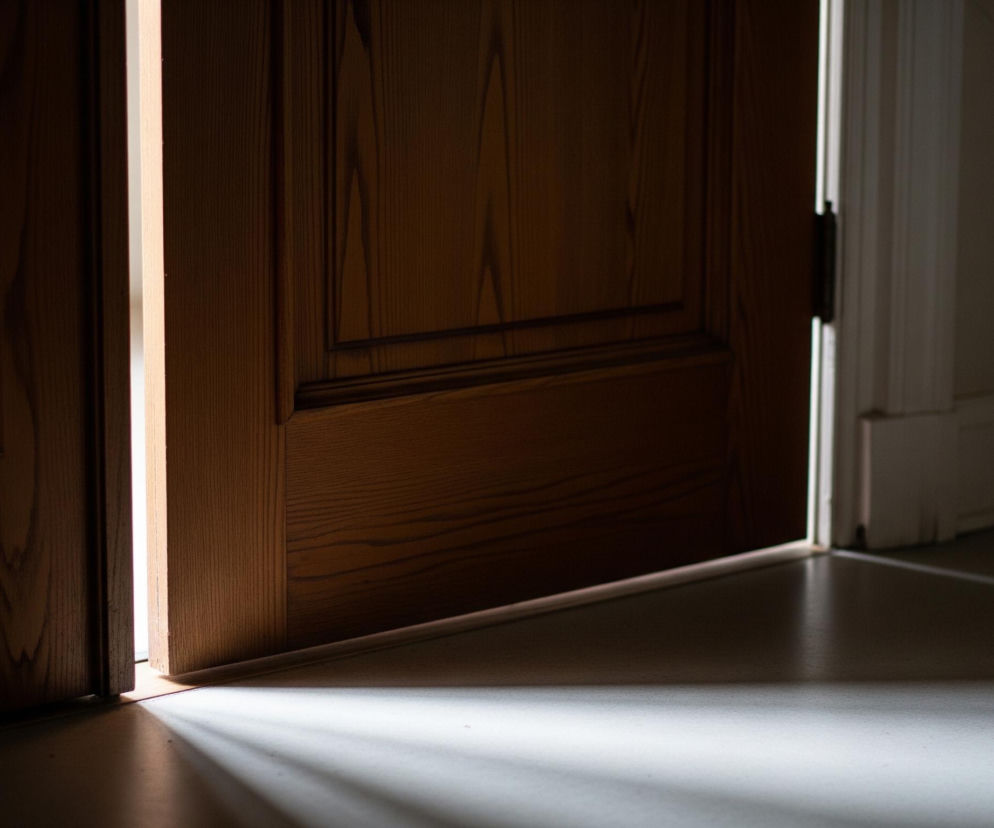 Uchylone drewniane drzwi z widocznym panelem ozdobnym na prawej części wypełniają niemal cały kadr, tworząc wąską szczelinę światła pośrodku. Zza tej szczeliny wydobywa się jasne, ciepłe światło, rzucające na podłogę promienie w kształcie trójkątów, które rozświetlają szarą powierzchnię podłoża. Po prawej stronie widoczny jest fragment białej futryny i ciemniejszej ściany. Dominują tu odcienie brązu i szarości, z kontrastującym jasnym akcentem.