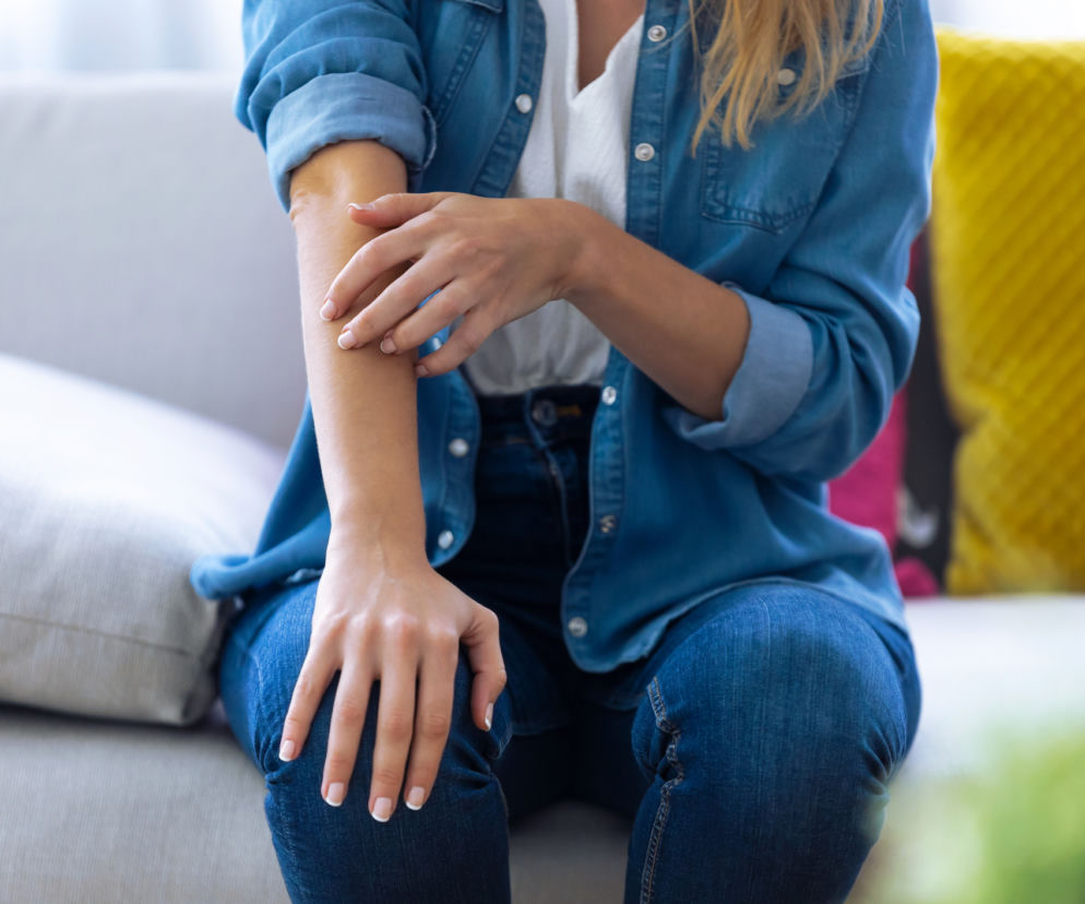 Blondynka w jeansowej koszuli i spodniach drapie się po przedramieniu, siedząc na jasnej kanapie z żółtą poduszką w tle. Zdjęcie ilustruje zjawisko zaraźliwego swędzenia, o którym można przeczytać na portalu Poradnik Zdrowie.