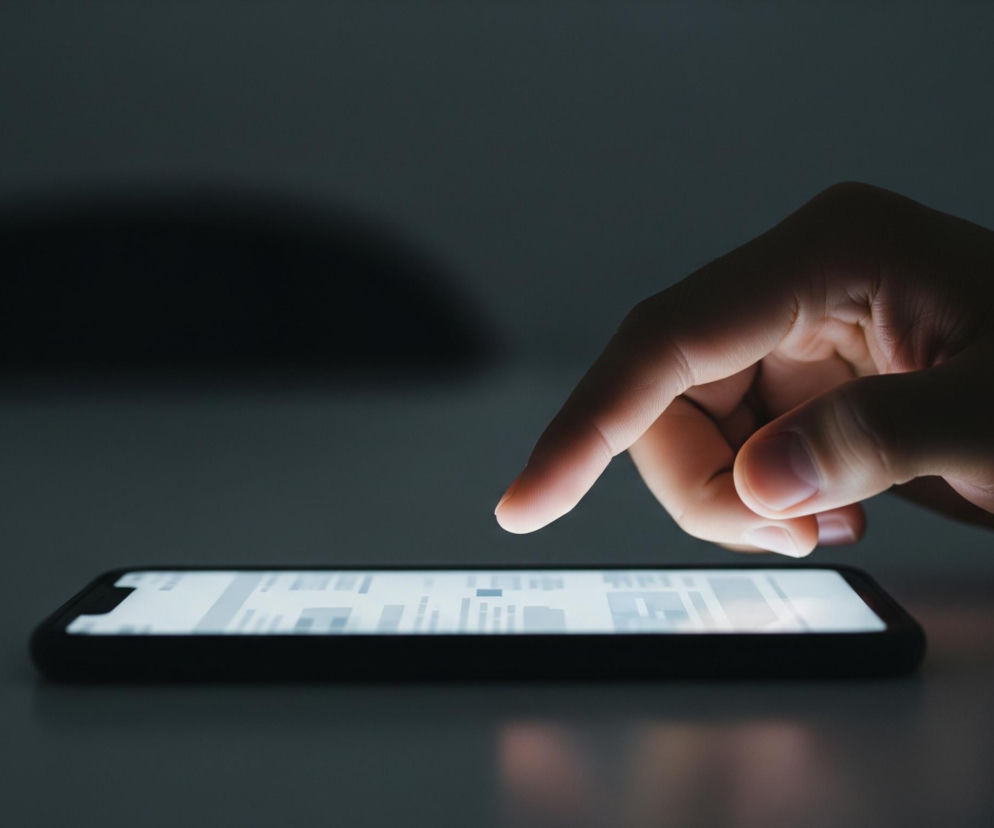Centralnie w kadrze znajduje się ludzka dłoń z palcem wskazującym dotykającym ekranu smartfona leżącego poziomo na ciemnej powierzchni. Ekran telefonu emituje jasne, białe światło, które rozjaśnia palec i część dłoni, tworząc subtelne odbicie na powierzchni pod urządzeniem. Reszta otoczenia jest pogrążona w ciemności, z ledwie widocznymi zarysami przedmiotów w tle, co podkreśla kontrast z jasnym ekranem.