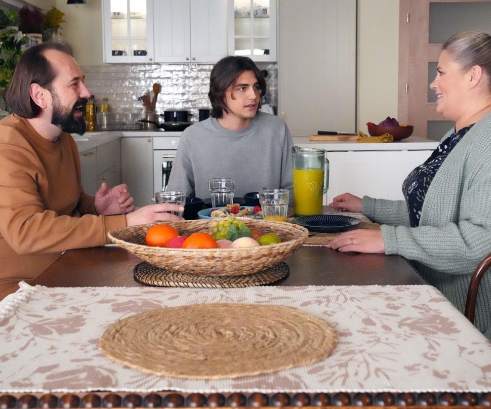 Rodzina Grzelaków, Aldona (Elżbieta Romanowska) i Borys (Jakub Wieczorek), siedzą przy stole z Romeo (Jacek Tyszkiewicz). Na stole miska z owocami i dzbanek z sokiem. Wszyscy rozmawiają, a uśmiechnięta Aldona patrzy na Romeo. Więcej o Barwach Szczęścia na Super Seriale.