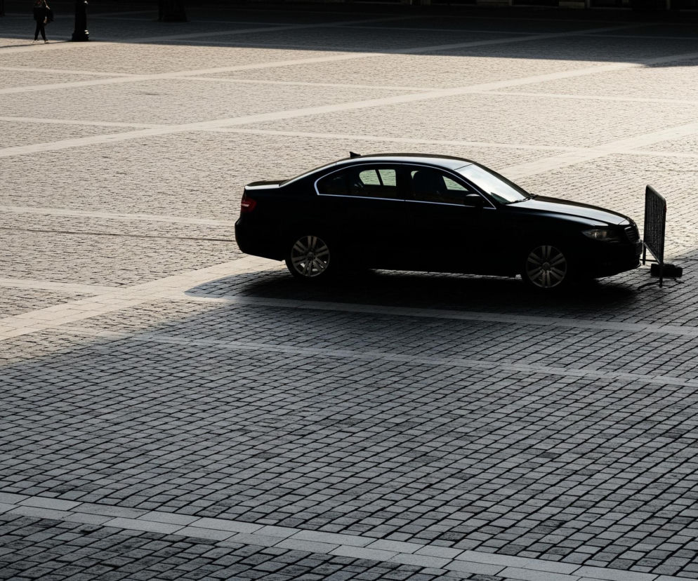 Na brukowanej, rozległej powierzchni widać czarny sedan zaparkowany na granicy światła i cienia. Przednie koło samochodu stoi na jasnym fragmencie nawierzchni, a reszta pojazdu jest w ciemnym cieniu. Przy przednim zderzaku samochodu po prawej stronie znajduje się metalowa barierka ochronna. Górna połowa obrazu jest jaśniejsza, ukazując oddalonych ludzi i zarys konstrukcji, natomiast dolna część pogrążona jest w ciemnym cieniu, co podkreśla kontrast na bruku.