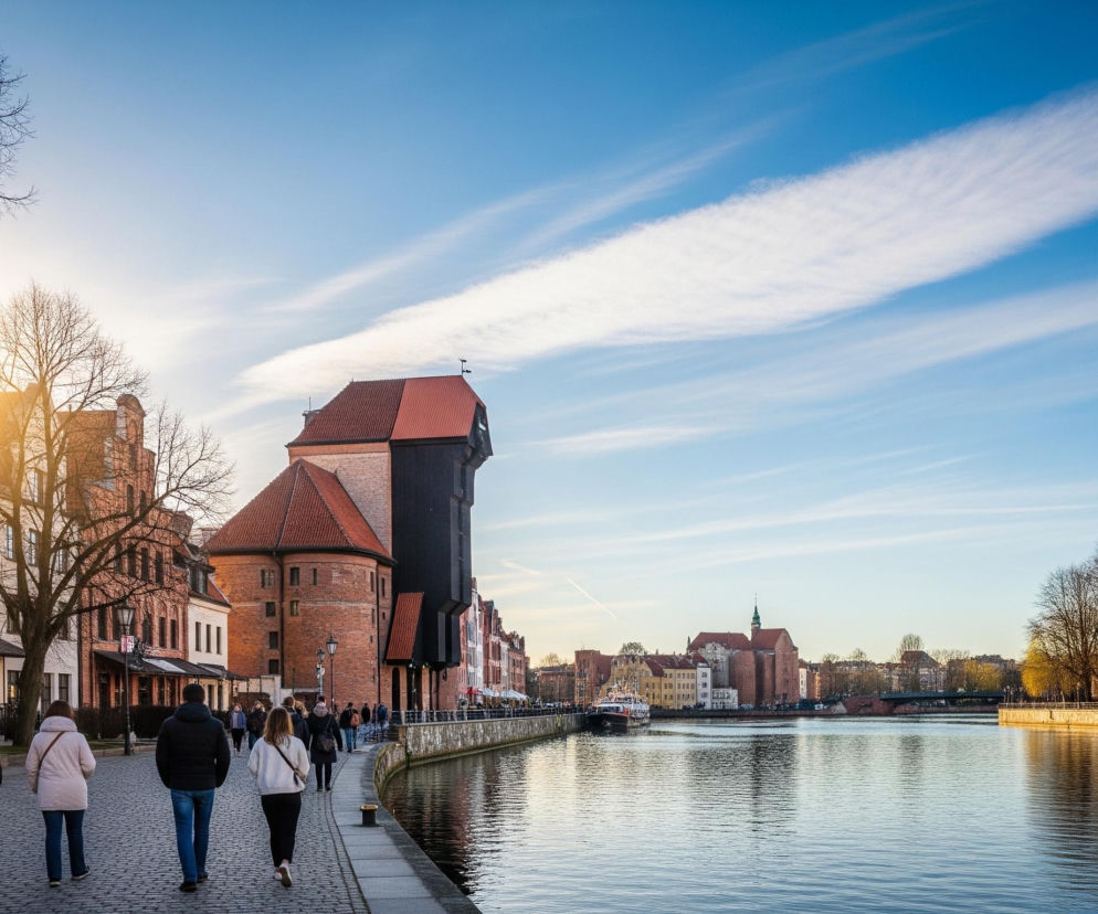 W kadrze dominuje szeroka perspektywa rzeki lub kanału, rozdzielająca dwie linie zabytkowej zabudowy i ludzi spacerujących brukowanym nabrzeżem. Po lewej stronie, w tle, wyróżnia się jasne słońce, które rozświetla fasady kamienic w pastelowych odcieniach, a ich sylwetki otaczają nagie drzewa. Centralny punkt stanowi duży, ceglany budynek z ciemnym drewnianym dźwigiem na dachu i czerwoną dachówką, przylegający do rzeki. Na niebie widać kilka białych, podłużnych chmur, ułożonych w paski, na tle intensywnie niebieskiego nieba.