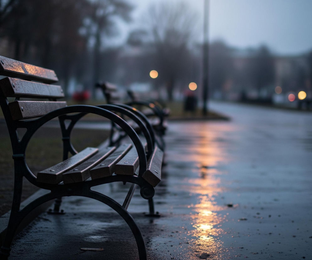 Na pierwszym planie, z lewej strony, widać mokrą drewnianą ławkę z czarnymi, metalowymi podłokietnikami i nogami. Ławka jest częściowo oświetlona pomarańczowo-żółtym światłem, odbijającym się na jej mokrej powierzchni. Przed ławką rozciąga się ciemny, mokry chodnik, na którym odbijają się rozmyte światła z oddali, tworząc długie, jasne smugi. W tle, po prawej stronie, widać rozmazaną ulicę z rzędem latarni, których światła są widoczne jako okrągłe, pomarańczowe i zielone plamy, a także zarysy budynków i drzew.