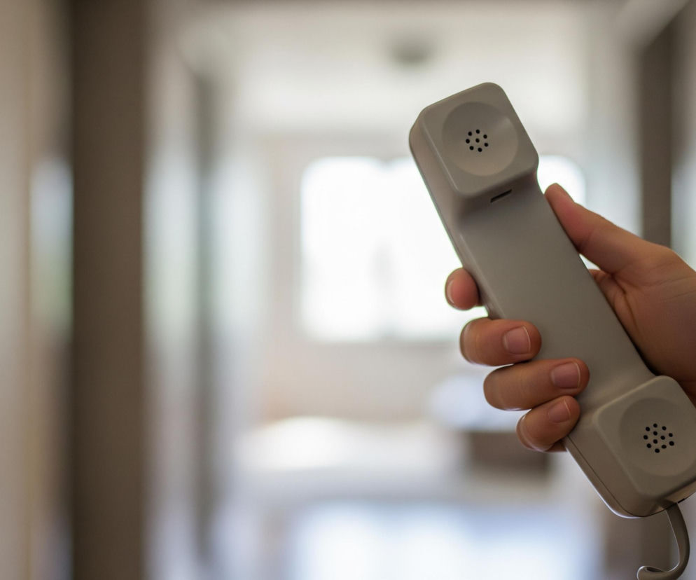 Dłoń trzyma beżową słuchawkę telefonu stacjonarnego, widoczną na pierwszym planie, skierowaną w stronę widza. Górna część słuchawki, z okrągłym zagłębieniem i sześcioma otworami głośnika, jest wyraźnie widoczna, podobnie jak dolna część z takimi samymi otworami mikrofonu i fragmentem zwiniętego kabla. Tło jest rozmyte, przedstawiając korytarz lub długie pomieszczenie z jasnym światłem w oddali, sugerującym okno lub otwarte drzwi.