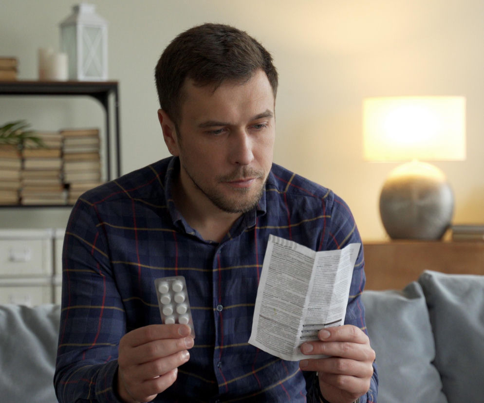 Mężczyzna w koszuli w kratę siedzi na kanapie, trzymając w jednej ręce blister z tabletkami, a w drugiej ulotkę, którą uważnie czyta. Wyraża zaniepokojenie dotyczące skutków ubocznych statyn. Więcej o ich bezpieczeństwie i efekcie nocebo przeczytasz na Poradnik Zdrowie.