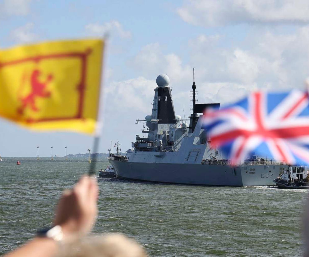 Brytyjski niszczyciel typu 45, D37 HMS Defender, płynący po morzu, obserwowany przez osoby z flagami Wielkiej Brytanii i Szkocji na pierwszym planie, w kontekście rewolucji obronności w Wielkiej Brytanii, o której możesz przeczytać na naszym portalu.