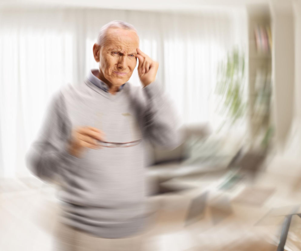 Starszy mężczyzna trzymający okulary w dłoni, z drugą ręką dotykającą skroni, wyraża ból głowy lub zawroty, co ilustruje temat artykułu. Rozmycie tła symbolizuje uczucie wirowania. Więcej o przyczynach zawrotów głowy przeczytasz na Poradnik Zdrowie.