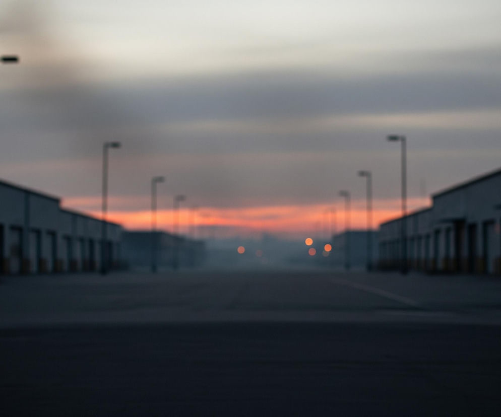 Rozmazaną perspektywę drogi biegnącej prosto, otoczonej z obu stron rzędami niskich, przemysłowych budynków. Na pierwszym planie po lewej i prawej stronie widoczne są latarnie uliczne o długich słupach i prostokątnych oprawach. Horyzont dominuje rozciągnięta chmura o barwach szarości i błękitu, z pomarańczowo-czerwonym akcentem w centrum, sugerującym wschód lub zachód słońca. W oddali, wzdłuż drogi, widać rozmyte pomarańczowe punkty świetlne.