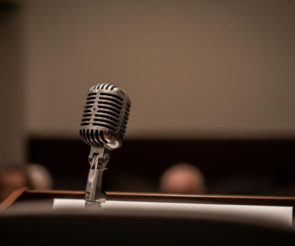 Centralnie umiejscowiony jest srebrny, retro mikrofon z kratką, stojący na ciemnobrązowej, drewnianej mównicy. Za mikrofonem widoczne są rozmyte, beżowo-brązowe kształty, sugerujące obecność innych osób lub elementów w tle. Całość kadru utrzymana jest w stonowanych, ciemnych barwach, z delikatnym oświetleniem padającym na mikrofon i górną część mównicy.