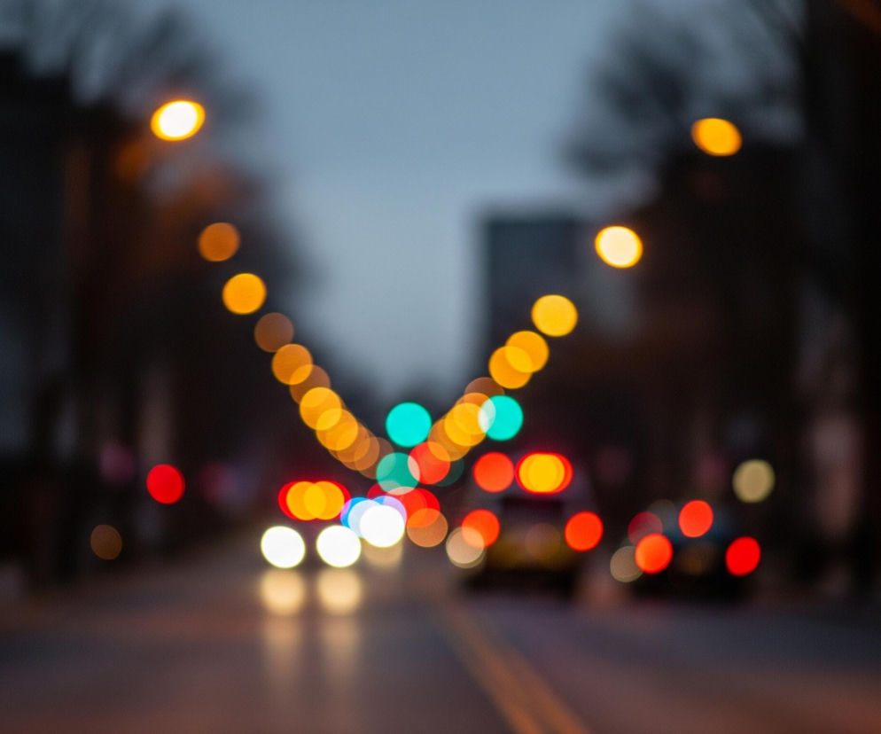 Rozmyta scena nocna przedstawia miejską ulicę z rzędami jasnych, kolistych świateł w różnych odcieniach. Na środku jezdni widoczne są dwie równoległe, żółte linie, zanikające w dali. Po obu stronach drogi rozmieszczone są pomarańczowe światła uliczne, tworzące łukowatą perspektywę, a bliżej środka obrazu znajdują się czerwone, białe i turkusowe plamy światła, sugerujące światła samochodów i sygnalizację świetlną. Tło stanowi zamazana sylwetka budynków i drzew pod ciemnym, granatowym niebem.