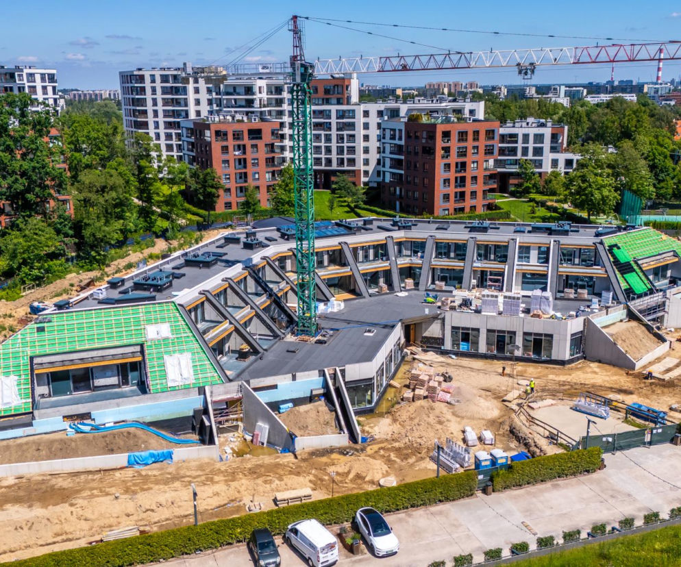 Widok z lotu ptaka na budowę luksusowego kompleksu mieszkaniowo-usługowego Rawelin VIII na warszawskim Ursynowie, z zielonymi dachami i żurawiem, otoczonego zielenią i nowoczesnymi blokami mieszkalnymi. Inwestycję opisywaną na portalu Architektura Murator Plus wyróżnia odtworzenie historycznej formy.