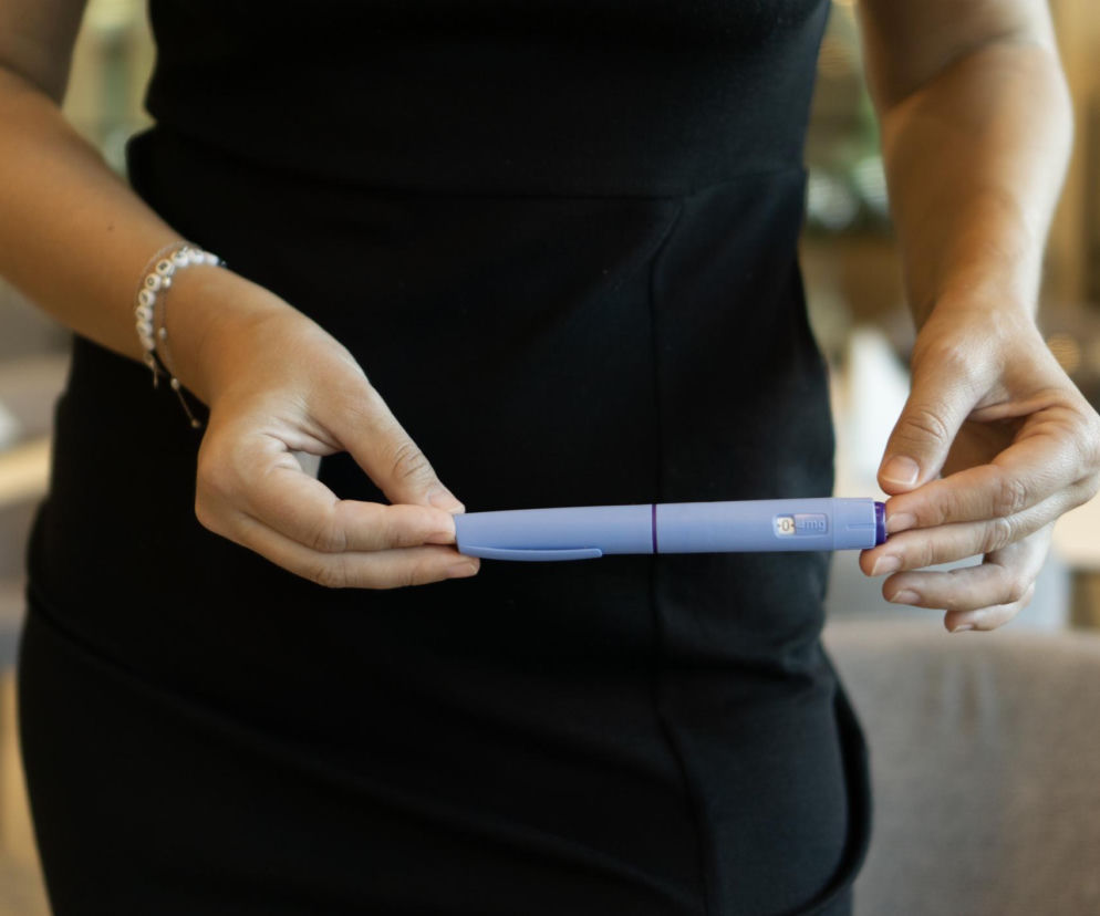 Kobieta w czarnej sukience trzyma fioletowy pen do wstrzykiwań z lekiem GLP-1 na odchudzanie, symbolizujący terapię. Zdjęcie ilustruje temat efektu jojo po lekach na odchudzanie, o którym można przeczytać na portalu Poradnik Zdrowie.