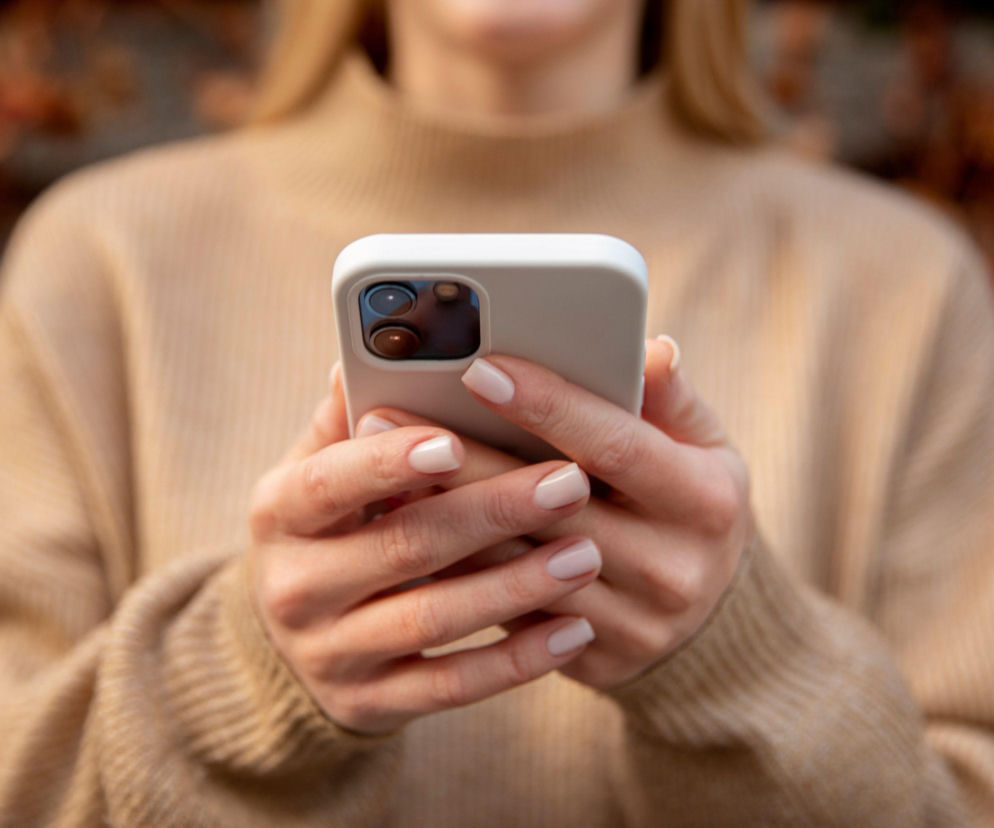 Dłonie kobiety trzymającej biały smartfon. O nowym systemie Alertu RCB przeczytasz w serwisie Eska.