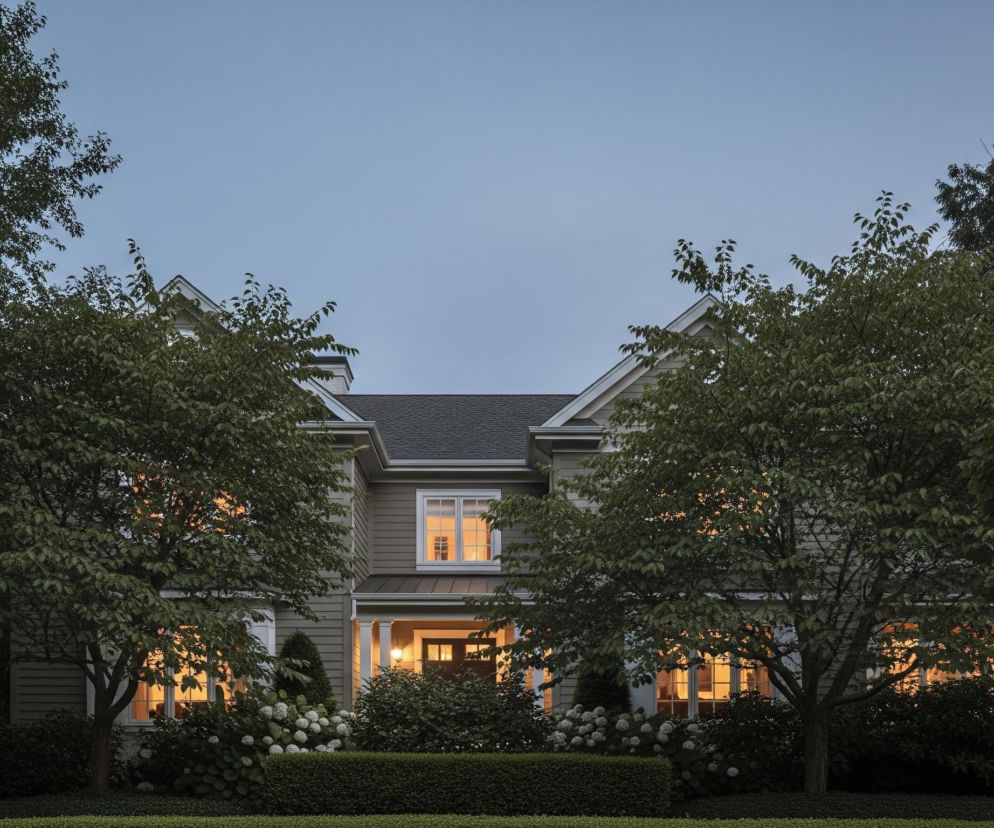 Pośrodku kadru znajduje się duży, dwupiętrowy dom w odcieniach szarości z ciemnym dachem i białymi obramowaniami okien oraz słupami werandy. W wielu oknach i na werandzie widać ciepłe, żółte światło, kontrastujące z niebieskawym, wieczornym niebem. Przed domem rośnie równo przycięty żywopłot oraz krzewy z białymi kwiatami, a po bokach kadru dominują gęste drzewa o zielonych liściach.