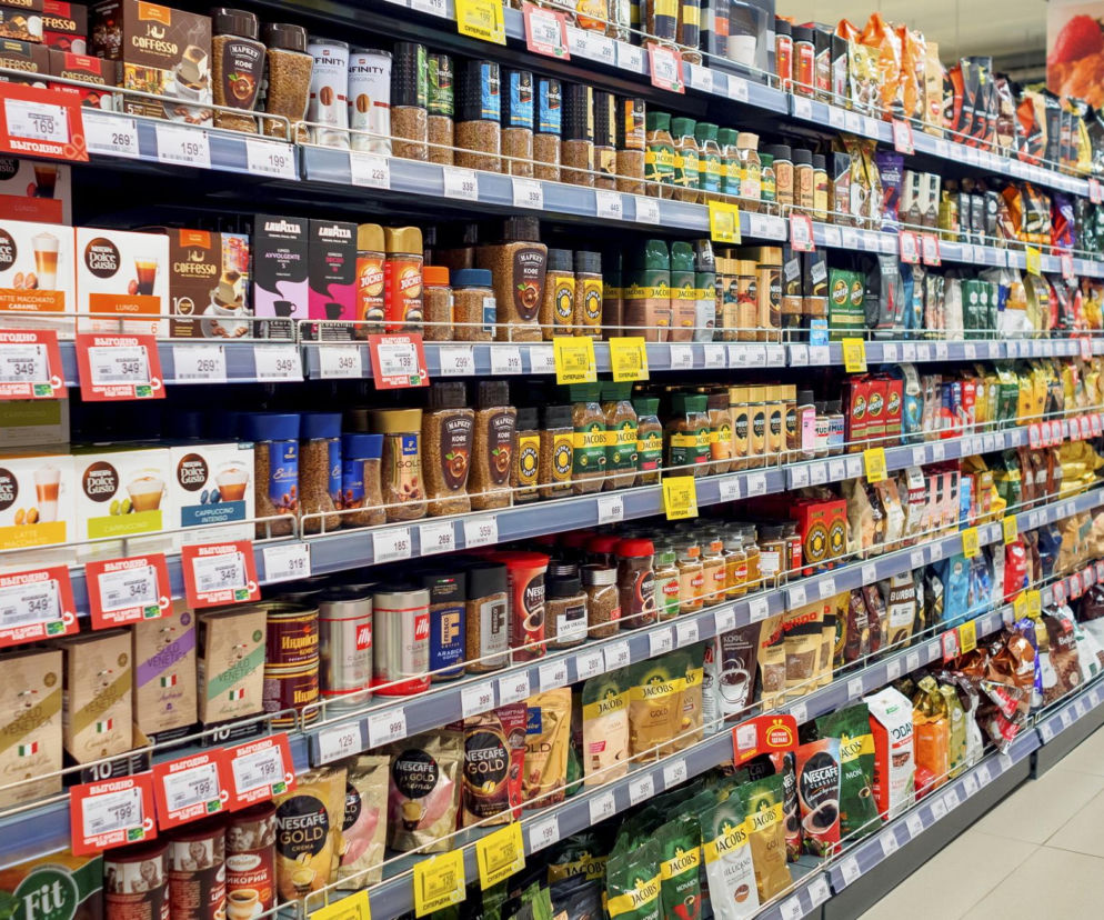 Regały w supermarkecie wypełnione różnymi opakowaniami kawy mielonej, rozpuszczalnej i w ziarnach, w tym markami jak Lavazza i Nescafe. Widoczne są też etykiety cenowe. Artykuł na Super Biznes informuje o wzroście cen kawy i podróbkach z domieszką soi.