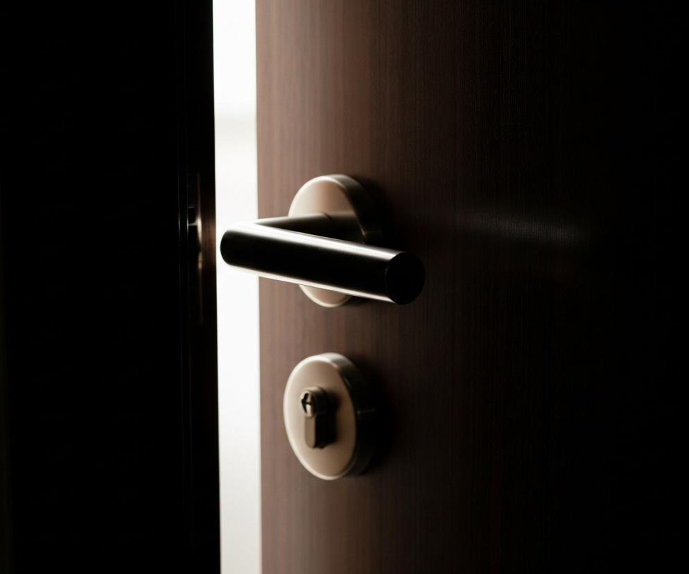 Uchylone drzwi ukazują jasną szczelinę światła pośrodku, między ciemnymi panelami. Po prawej stronie, na powierzchni drzwi o fakturze drewna w kolorze ciemnego brązu, widoczne są dwa metalowe elementy: klamka o cylindrycznym kształcie i podłużny zamek z okrągłą tarczą. Lewa strona obrazu to całkowicie czarna przestrzeń, która przechodzi w ciemny brzeg framugi przy szczelinie światła.