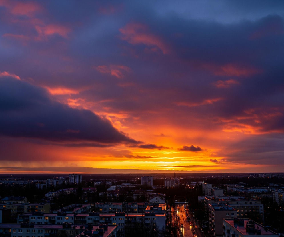 Miasto o zachodzie słońca skąpane jest w intensywnych barwach, z pomarańczowo-czerwonym niebem dominującym w centrum kadru. Chmury rozciągają się od dolnej krawędzi obrazu, gdzie przyjmują ciemne, fioletowo-niebieskie odcienie, aż po górną część, gdzie ich brzegi są podświetlone intensywną pomarańczą. Poniżej horyzontu rozciąga się miejski krajobraz z licznymi budynkami mieszkalnymi, których dachy i fasady są częściowo widoczne w ciemnych, stonowanych barwach. Na ulicach widać odbicia świateł, a kilka samochodów z włączonymi reflektorami porusza się po mokrej nawierzchni.