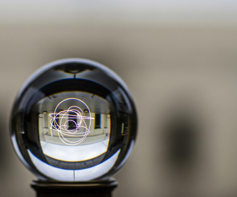 W centrum kadru znajduje się przezroczysta szklana kula, odbijająca i odwracająca otoczenie. Wewnątrz kuli widoczne są jasno świecące, nieregularne linie, przypominające spirale lub zwoje, o fioletowo-białym zabarwieniu. Odbicie ukazuje również fragmenty budynku z pionowymi, ciemnymi oknami i jasnymi ścianami, z odwróconą perspektywą. Kula spoczywa na ciemnym, cylindrycznym cokole, a tło jest jednolite, rozmyte, o jasnobrązowo-szarym kolorze.