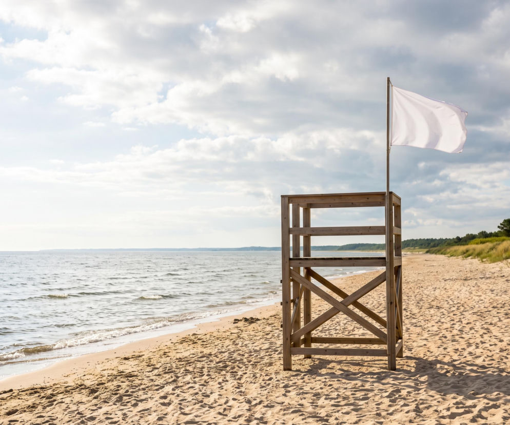 Drewniana wieża ratownika z białą flagą na pustej, piaszczystej plaży. O pogodzie w Chłopach przeczytasz na SE Szczecin.