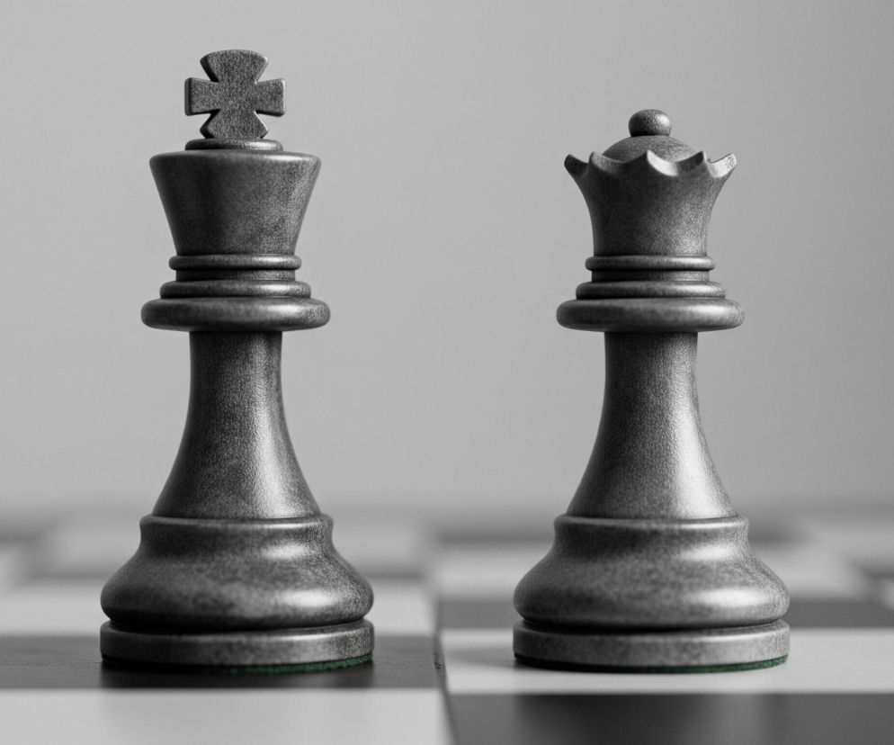 Na zdjęciu widoczne są dwie figurki szachowe: król i królowa, ustawione obok siebie na szachownicy. Obie figurki są ciemnoszare z widoczną teksturą przypominającą kamień lub metal, a ich podstawy mają cienkie zielone podkładki. Król, po lewej stronie, jest wyższy, zakończony krzyżem, natomiast królowa po prawej ma koronę z ząbkowanym brzegiem i małą kulkę na szczycie. Tło stanowi rozmyta szachownica z białymi i czarnymi polami, przechodząca w jasnoszary, jednolity kolor w górnej części obrazu.