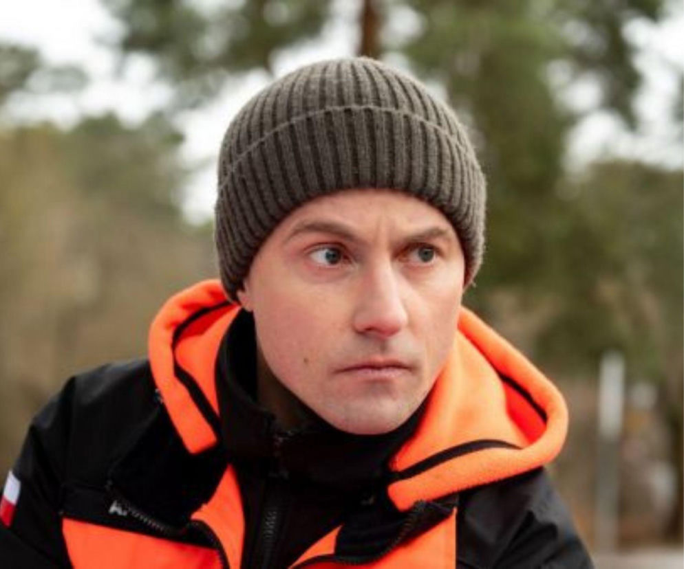 Mężczyzna w szarej czapce i pomarańczowo-czarnej kurtce ratowniczej z polską flagą na ramieniu, patrzący w prawą stronę, w tle rozmazane drzewa. Obraz ten ilustruje wydarzenia z 886. odcinka serialu Na sygnale, o którym można przeczytać więcej na portalu Super Seriale.