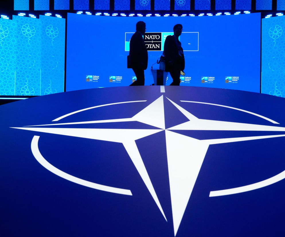 Logo NATO na niebieskim tle. W głębi sylwetki osób na forum. O inwestycjach Sojuszu przeczytasz na SE Portal Obronny.
