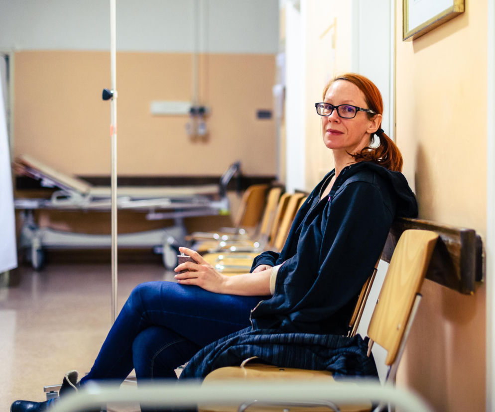 Kobieta z rudymi włosami w okularach, siedząca na drewnianej ławce w szpitalnym korytarzu, symbolizująca problem długich kolejek na badania. W tle widoczne szpitalne łóżka. Na portalu Poradnik Zdrowie znajdziesz informacje o nowych limitach NFZ.