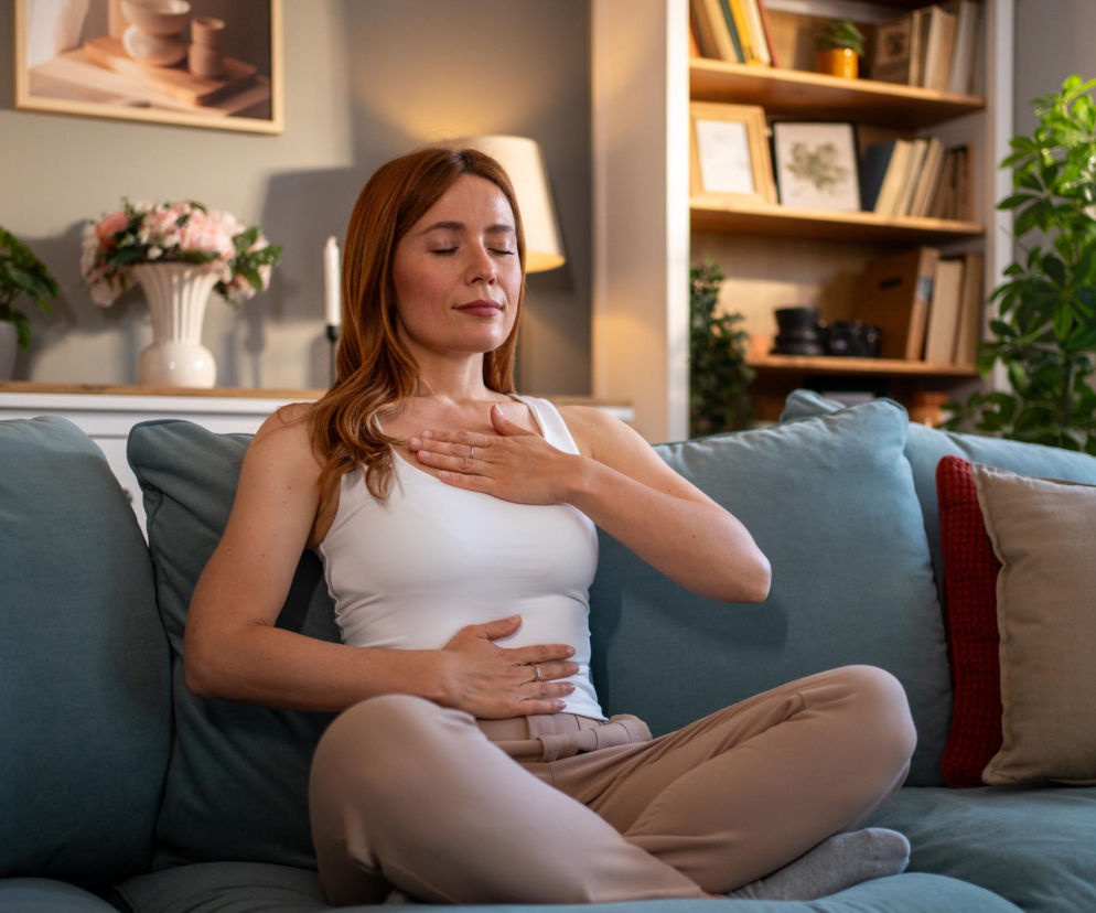 Kobieta o rudych włosach, ubrana w białą koszulkę i beżowe spodnie, siedzi w pozycji medytacyjnej na niebieskiej kanapie, dotykając dłonią klatki piersiowej i brzucha. W tle domowe, spokojne wnętrze z półkami na książki i roślinami. Zdjęcie ilustruje techniki oddechowe, o których można przeczytać na portalu Poradnik Zdrowie.