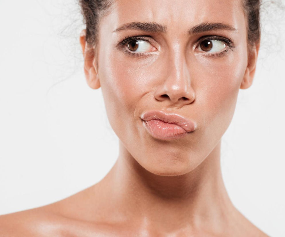 Zbliżenie na twarz kobiety, która marszczy nos i przymyka usta, patrząc w bok z wyrazem lekkiego zamyślenia lub niepewności, na tle jasnego tła. Takie gesty mogą świadczyć o nieprzyjemnym problemie pękających kącików ust. Więcej o zajadach dowiesz się na Poradnik Zdrowie.