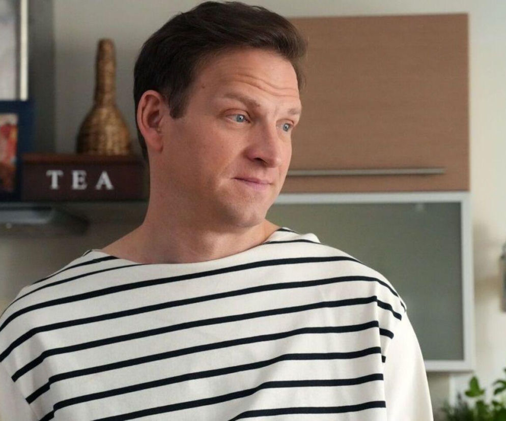 Łukasz (Michał Rolnicki) z serialu Barwy szczęścia w marynarskim swetrze, z delikatnym uśmiechem i zamyślonym wyrazem twarzy, patrzy w prawo. Tło przedstawia fragment kuchni z szafkami i pudełkiem na herbatę. Więcej o jego kłopotach znajdziesz na Super Seriale.