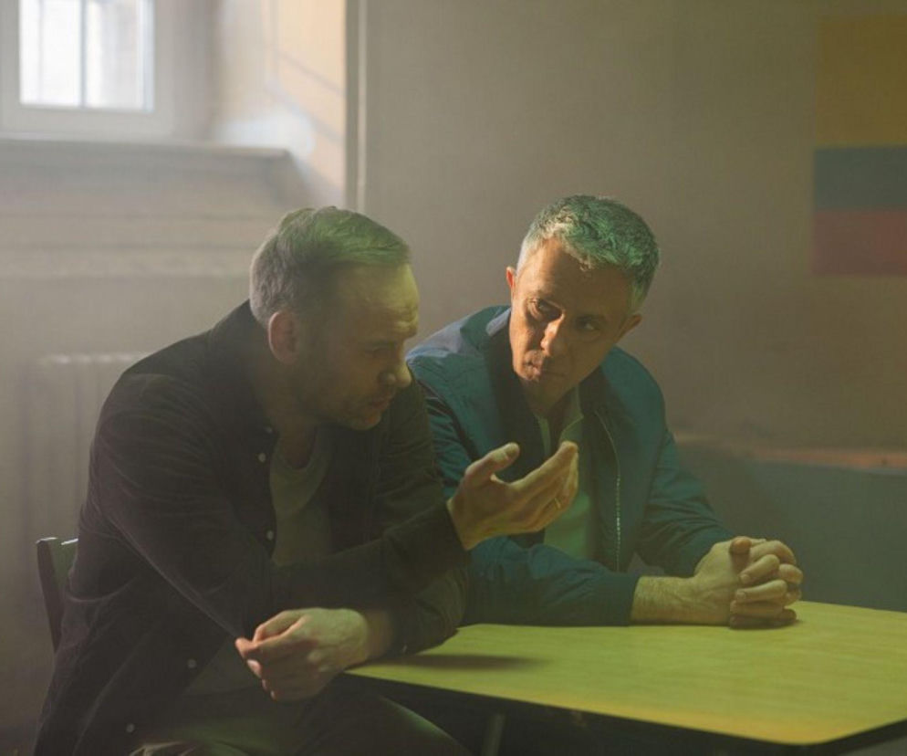 Janek i Artur, dwaj mężczyźni o zmartwionych minach, siedzą przy stole i rozmawiają w tajemniczym, lekko zadymionym pomieszczeniu. W tle widoczna flaga Kolumbii. Scena ta odzwierciedla napięcie z odcinka 4189 Pierwszej miłości, którą można oglądać na Super Seriale.