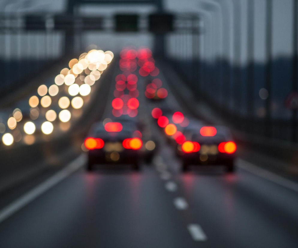 Rozmyty widok przedstawia autostradę o zmierzchu lub w nocy, z rzędami jasnych świateł pojazdów. Dwa wyraźniejsze światła tylne samochodów w kolorze czerwono-pomarańczowym znajdują się na prawym pasie, a przed nimi widoczny jest długi sznur rozmytych czerwonych świateł. Na lewym pasie widoczne są rozmyte, okrągłe, jasne światła przednie pojazdów. Droga jest ciemnoszara, a na niej widać białe, przerywane linie, dzielące pasy ruchu. W tle widać ciemne, rozmyte konstrukcje mostu oraz zarys linii drzew.