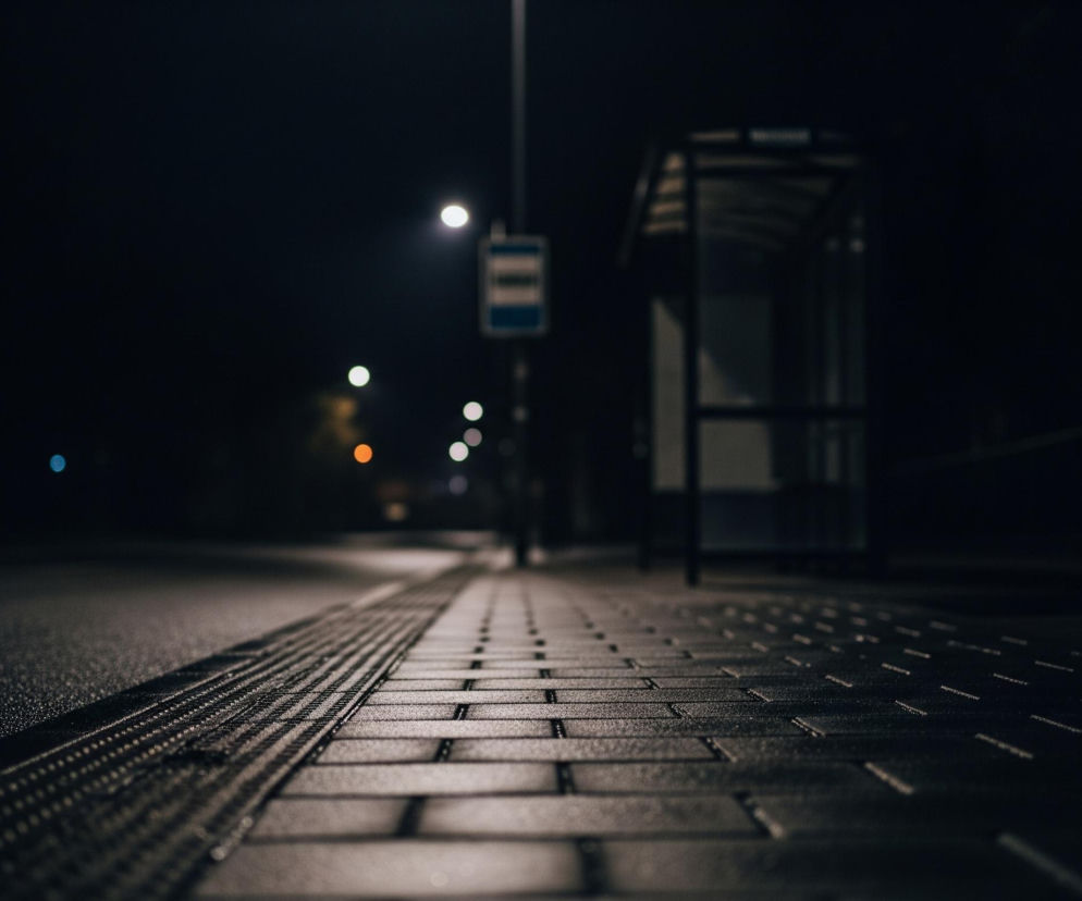 Nocna sceneria przedstawia chodnik z ciemnych, prostokątnych płyt ułożonych w jodełkę, przechodzący w asfaltową drogę po lewej stronie. Wzdłuż krawędzi chodnika biegnie pas ostrzegawczy z wypustkami, odbijający światło. Po prawej stronie w oddali znajduje się rozmyty przystanek autobusowy z przezroczystymi ściankami i ławką. Centralnie umieszczona latarnia uliczna oświetla scenę, a w tle widać rozmyte, jasne punkty świateł.