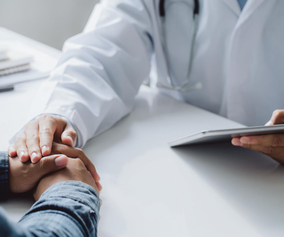 Lekarz w białym kitlu z ręką pocieszająco położoną na dłoniach pacjenta w koszuli w kratę. Na stole notatnik i tablet. Zdjęcie symbolizuje wsparcie w leczeniu HIV, o którym przeczytasz więcej na Poradnik Zdrowie.