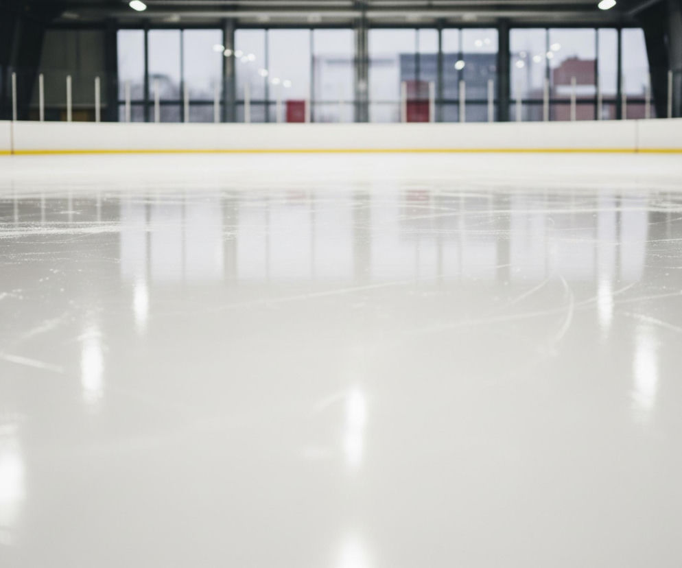 Lód na arenie sportowej pokrywa cały pierwszy plan i większość środkowego planu obrazu, ukazując liczne smugi i drobne cząsteczki lodu, a także odbicia świateł sufitowych. W tle rozciąga się biała banda okalająca lodowisko, z wyraźną żółtą linią na dole, a wzdłuż niej widoczne są krótkie pionowe pasy w kolorach niebieskim i czerwonym. Za bandą znajduje się przeszklona ściana, przez którą widać zarysy budynków zewnętrznych oraz odbicia świateł, co tworzy rozmyty, miejski krajobraz.