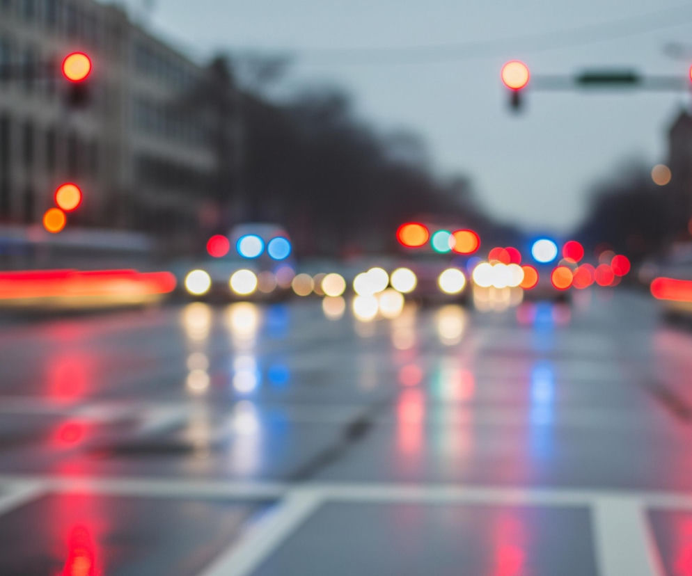 Rozmyte ujęcie miejskiej ulicy o zmierzchu lub w nocy, z widocznymi światłami samochodów i sygnalizacji świetlnej. Dominują kolory czerwony, pomarańczowy i niebieski, pochodzące od źródeł światła, które rozpraszają się na mokrej nawierzchni jezdni. Na pierwszym planie widać szarą jezdnię z białymi, rozmytymi pasami, a w tle zarys budynków i drzew. Światła tworzą podłużne smugi i okrągłe plamy, odzwierciedlając się w błyszczącej powierzchni drogi.