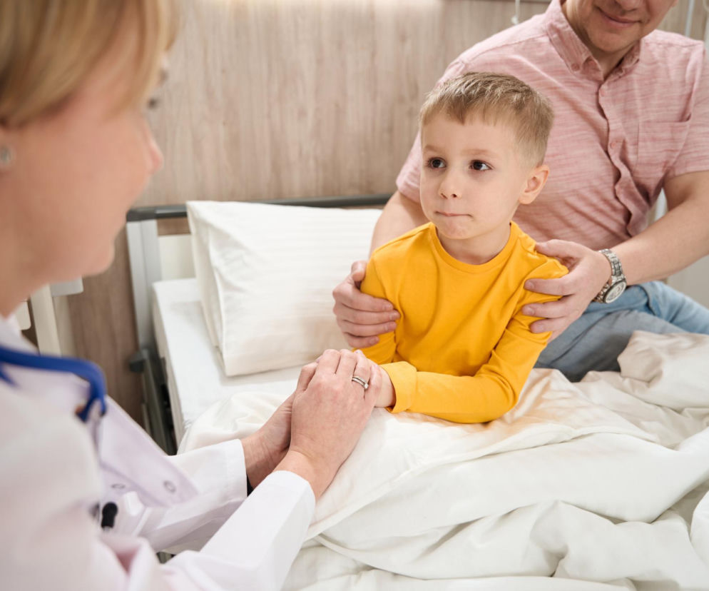 Lekarka w białym kitlu ze stetoskopem trzyma za ręce siedzącego w łóżku małego chłopca w żółtej bluzce, podczas gdy dorosły mężczyzna w różowej koszuli trzyma chłopca za ramiona, wspierając go. O wczesnych objawach nowotworów dziecięcych przeczytasz na Poradnik Zdrowie.