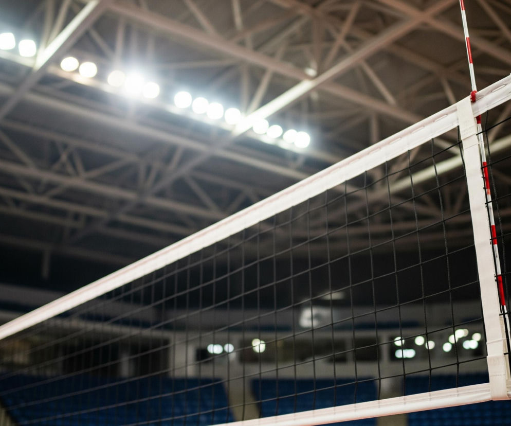 Siatka sportowa rozpięta na tle słabo oświetlonej hali. Górna krawędź siatki jest biała i grubsza, natomiast reszta siatki składa się z czarnych, równomiernie splecionych oczek. Po prawej stronie widoczny jest pionowy słupek w biało-czerwone pasy, przymocowany do siatki oraz element mocujący siatkę. W tle majaczą zarysy trybun z granatowymi siedzeniami, a ponad nimi widoczne są liczne jaskrawo świecące światła na suficie.