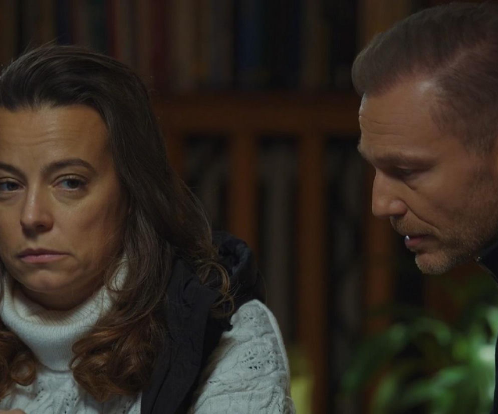 Magda Budzyńska (Anna Mucha) w białym, dzianinowym golfie z czarną kamizelką, z długimi, brązowymi włosami i smutnym wyrazem twarzy, obok niej Andrzej Budzyński (Krystian Wieczorek) z profilu. Para z serialu M jak miłość decyduje o przyszłości fundacji, o czym przeczytasz na Super Seriale.