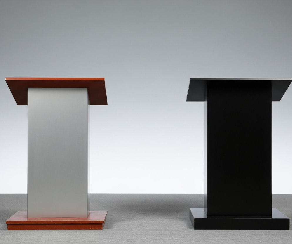 Dwa proste, prostopadłościenne postumenty stoją na jasnoszarej wykładzinie. Ten po lewej stronie ma szary korpus, zwieńczony ciemnoczerwonym blatem i oparty na takiej samej ciemnoczerwonej podstawie. Postument po prawej jest całkowicie czarny, z czarnym blatem i podstawą. Oba obiekty są oświetlone, a za nimi rozciąga się jaśniejsze, neutralne tło, przechodzące od jasnoszarego do białego.