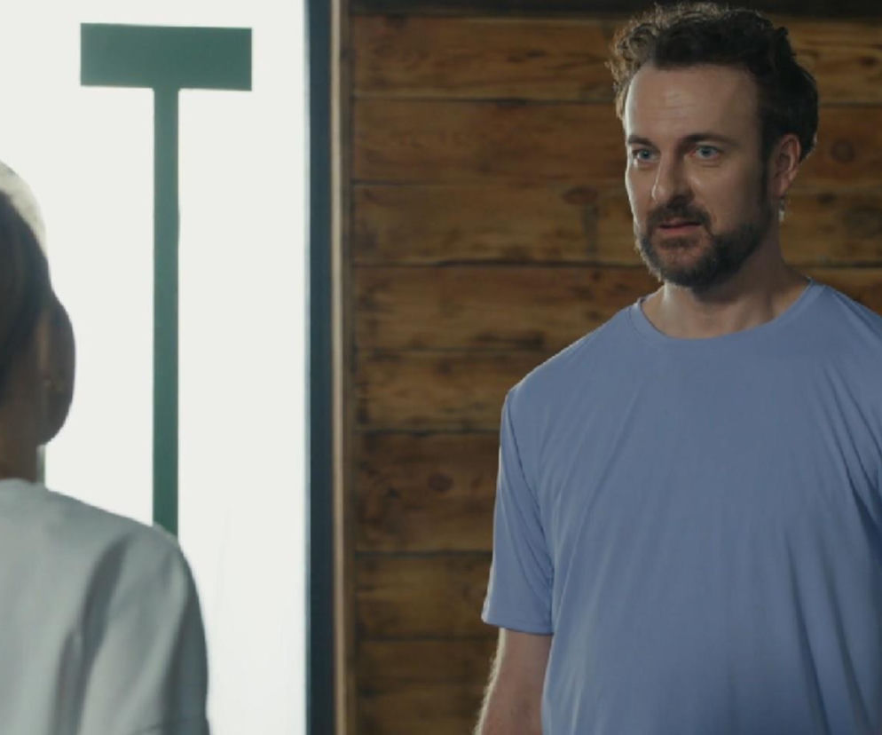 Radomir (Łukasz Wójcik) z serialu Barwy szczęścia w niebieskiej koszulce z krótkim rękawem, patrzący z niepokojem na kobietę w jasnym stroju, której widzimy tył głowy. Scena po wspólnej nocy z Dorotą, gdzie Radomir obawia się konsekwencji zdrady żony. Więcej o losach bohatera przeczytasz na Super Seriale.