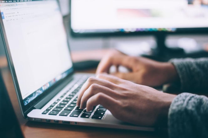 Dłonie osoby w szarym swetrze obsługujące laptopa z otwartym programem do fakturowania, w tle rozmyty ekran monitora. Zdjęcie ilustruje codzienną pracę biurową i wybór odpowiedniego programu do fakturowania, o czym więcej przeczytasz na naszym portalu.