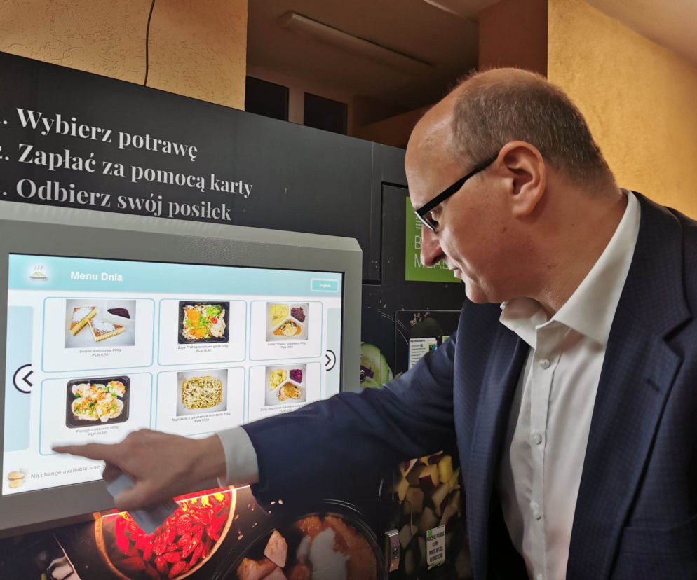Mężczyzna w okularach i garniturze wybiera danie z ekranu dotykowego automatu vendingowego oferującego ciepłe posiłki. Obok ekranu instrukcje wyboru i płatności kartą. To nawiązanie do dyskusji o kosztach kas fiskalnych w automatach, o czym piszemy na naszym portalu.
