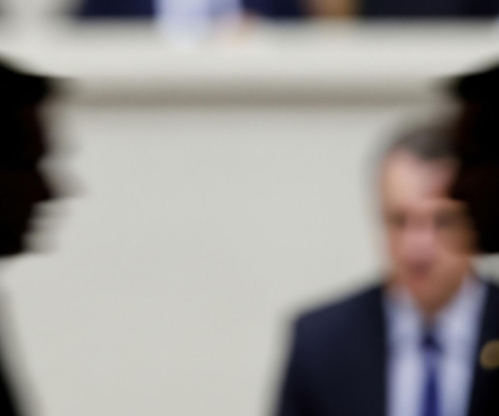 Na pierwszym planie, po lewej i prawej stronie, znajdują się rozmazane sylwetki dwóch mężczyzn w ciemnych garniturach. Ich profile są widoczne, a twarze i reszta ciała zlewają się w ciemne, nieostre kształty. Pomiędzy nimi, w centrum obrazu, widać nieostrą postać trzeciego mężczyzny, również w ciemnym garniturze i jasnej koszuli z niebieskim krawatem, z zamazaną, jasną twarzą. Tło jest jednolicie beżowe lub jasnoszare, całkowicie rozmyte.