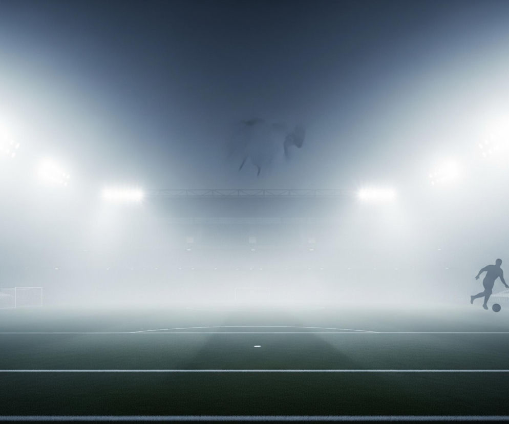 Płyta boiska piłkarskiego wypełnia dolną część kadru, z wyraźnie widocznymi białymi liniami oznaczającymi pole gry na zielonej murawie. Stadion oświetlony jest przez silne reflektory, które tworzą gęstą mgłę lub dym, intensywnie rozświetlając całą przestrzeń i sprawiając, że trybuny w tle są ledwo widoczne. Sylwetka piłkarza dryblującego piłkę znajduje się w prawej części kadru, ciemna i kontrastująca z jasnym tłem. W górnej, centralnej części kadru niewyraźnie zarysowują się zarysy dwóch postaci na tle mgły.
