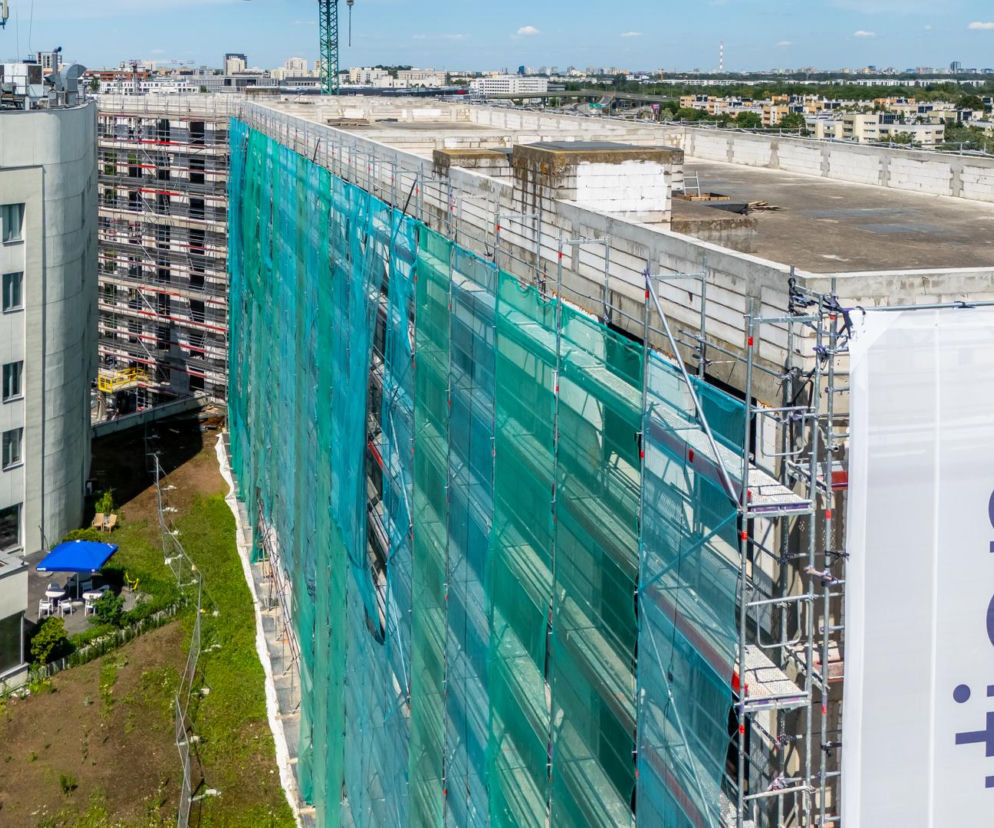 Budowa hotelu Holiday Inn w Warszawie przy Sobieskiego, gdzie szkieletor przekształca się w nowoczesny obiekt. Elewacja z rusztowaniami i siatką ochronną, symbolizująca rewitalizację w Architektura Murator Plus.