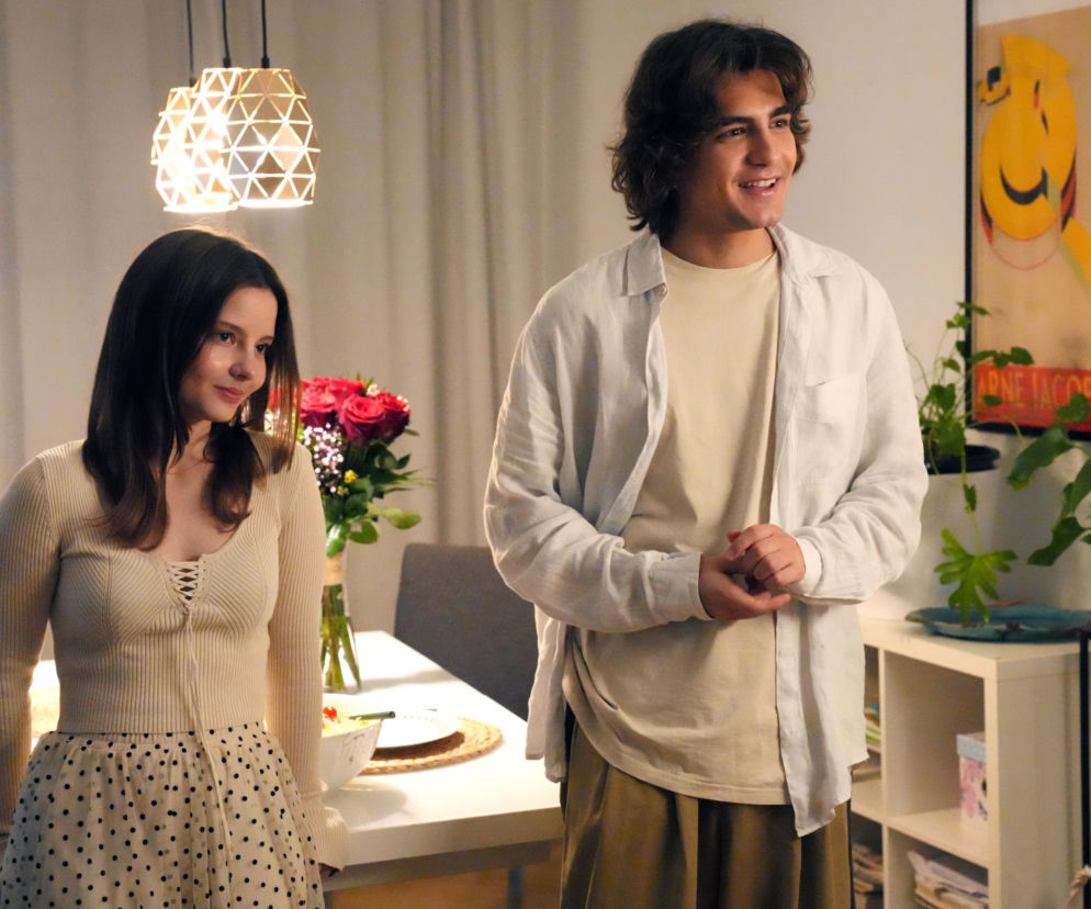 Ewa (Gabriela Ziembicka) i Romeo (Jacek Tyszkiewicz) w scenie z serialu Barwy szczęścia, stojący w pokoju, uśmiechnięci, po lewej bukiet czerwonych róż na stole. O ich losach przeczytasz na portalu Super Seriale.