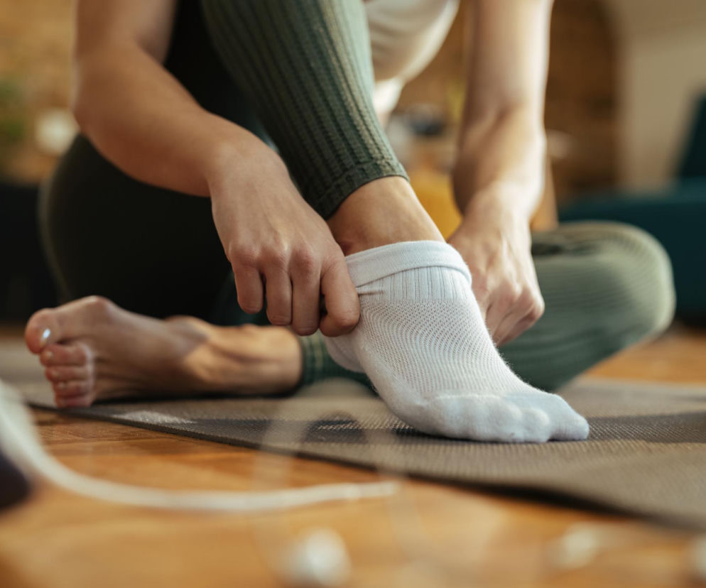 Kobieta w zielonych legginsach siedzi na macie do ćwiczeń na drewnianej podłodze, ściągając białą skarpetkę z prawej stopy, ukazując lewą bosą stopę z pomalowanymi paznokciami. To obraz problemów z krążeniem, o których przeczytasz na Poradnik Zdrowie.
