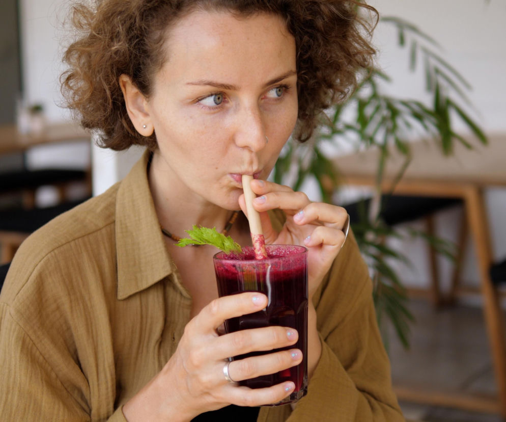 Kobieta z kręconymi włosami w beżowej koszuli pije ciemnoczerwony smoothie przez słomkę z bambusa, z listkiem selera jako dekoracją. W tle rozmazane stoliki i krzesła w kawiarni. Zdjęcie ilustruje zdrowe nawyki żywieniowe wspierające serce, o których przeczytasz na portalu Poradnik Zdrowie.