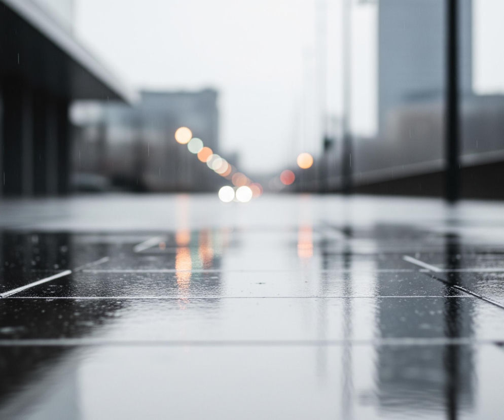 Mokra, ciemna, kwadratowa nawierzchnia z płyt chodnikowych dominuje dolną część kadru, odbijając rozmazane, jasne kolory świateł w oddali. Na powierzchni widać pojedyncze krople wody i drobne fale, świadczące o padającym deszczu. W tle, rozmyte kontury budynków miejskich tworzą industrialny pejzaż, natomiast horyzont rozświetlają okrągłe, nieostre światła w barwach pomarańczowych, białych i zielonkawych. Całość utrzymana jest w chłodnej palecie szarości i błękitów, z akcentami ciepłych barw świateł.